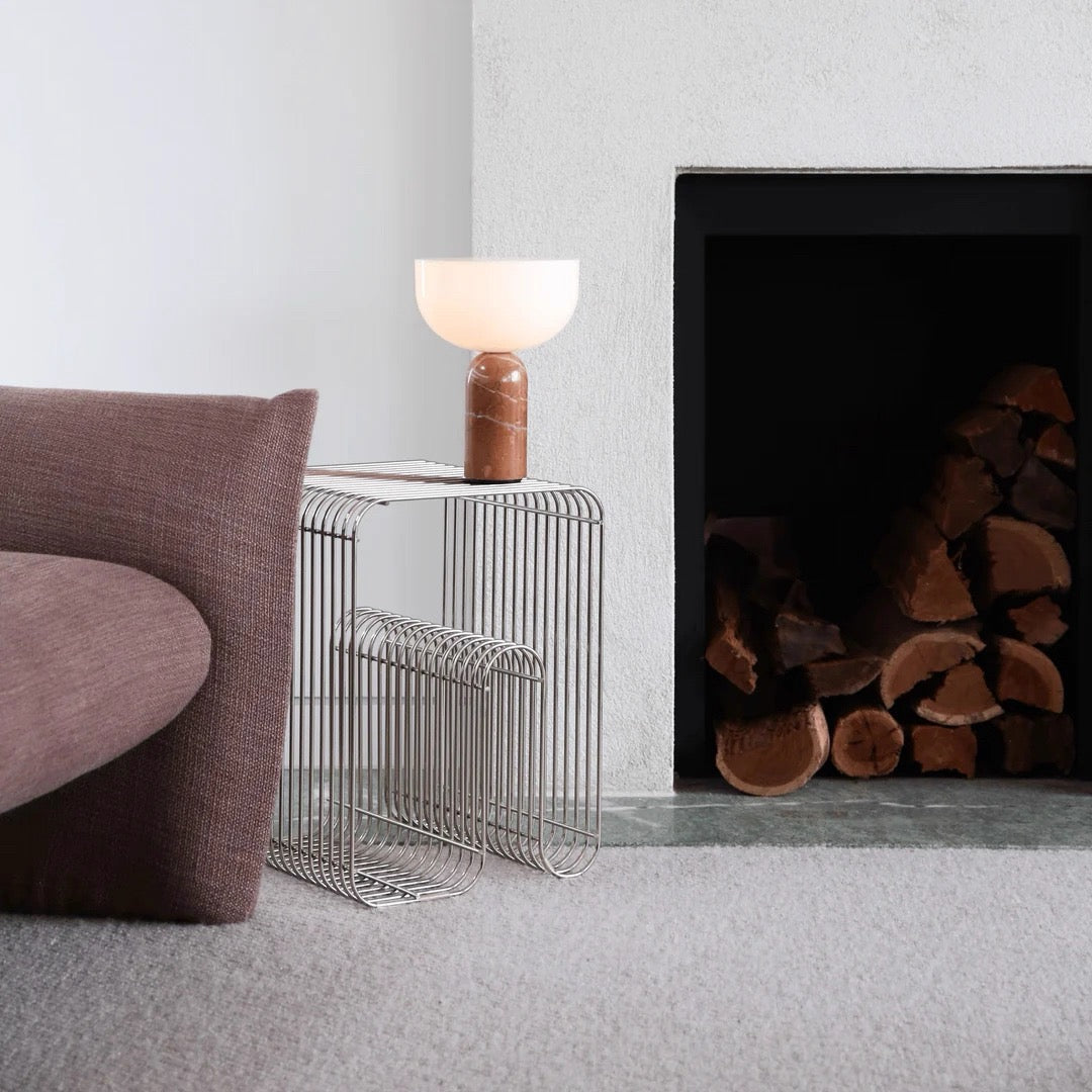Modern side table with a lamp next to a fireplace with wood logs.