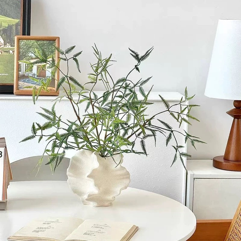 Decorative plant in a white vase on a table with a lamp and books in the background.