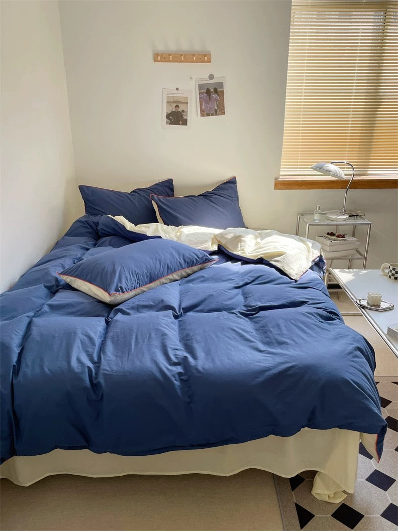 Bedroom with a bed covered in blue bedding and pillows, nightstand with lamp, and window with blinds.