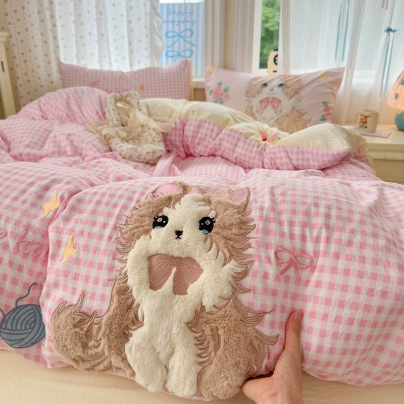 Hand holding a plush toy with a cat design on a bed with pink checkered bedding.