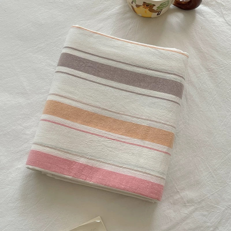 A folded white cotton muslin swaddle blanket with horizontal stripes in dusty pink, peach, mauve, and gray, styled on a white textured fabric surface.