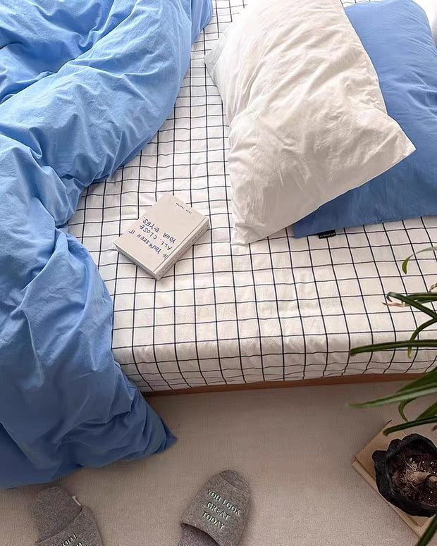 Blue bedding on a bed with a grid-patterned sheet, white pillow, and gray slippers on the floor.