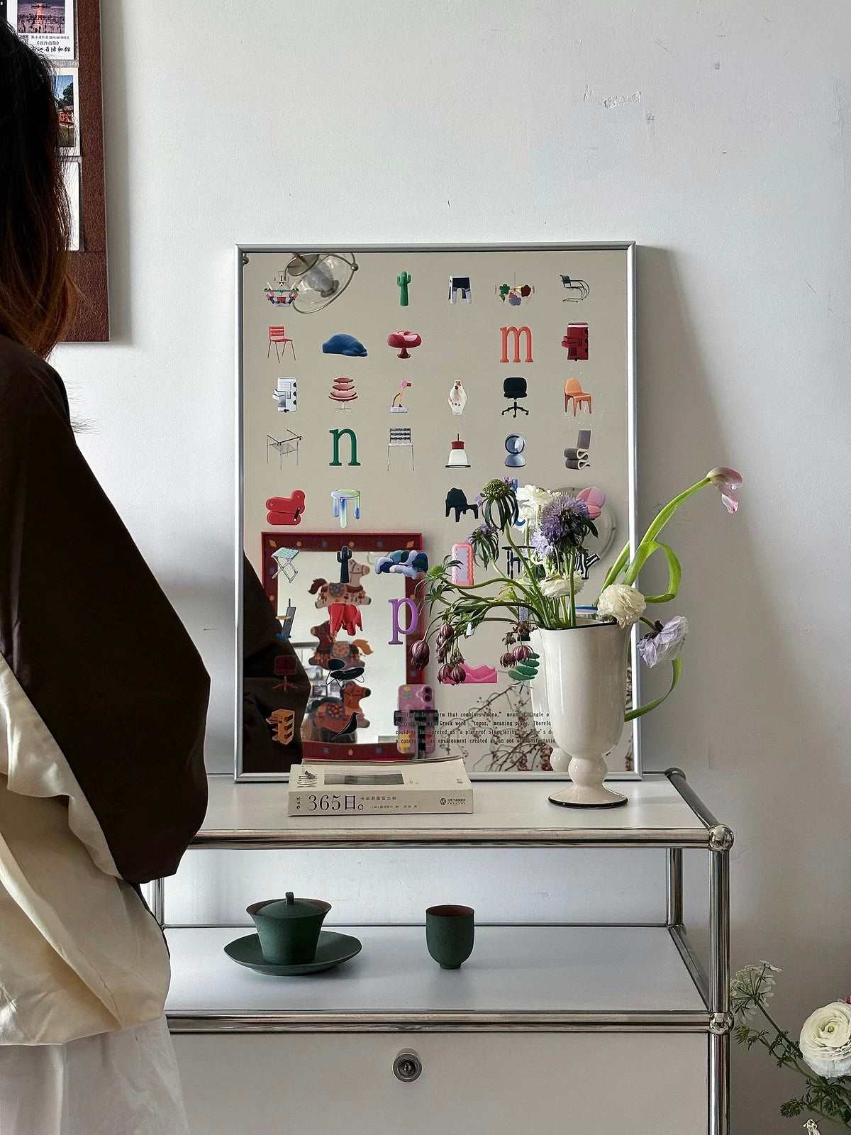 A framed art print featuring a collage of small furniture icons and letters, displayed on a white shelf in a styled room next to a person.