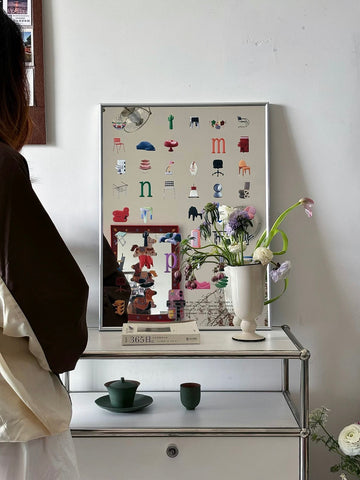 A framed art print featuring a collage of small furniture icons and letters, displayed on a white shelf in a styled room next to a person.