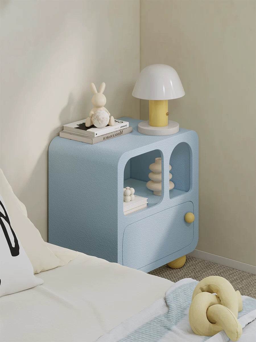 A blue arched nightstand beside a bed has yellow ball feet, a yellow knob and two open display compartments.