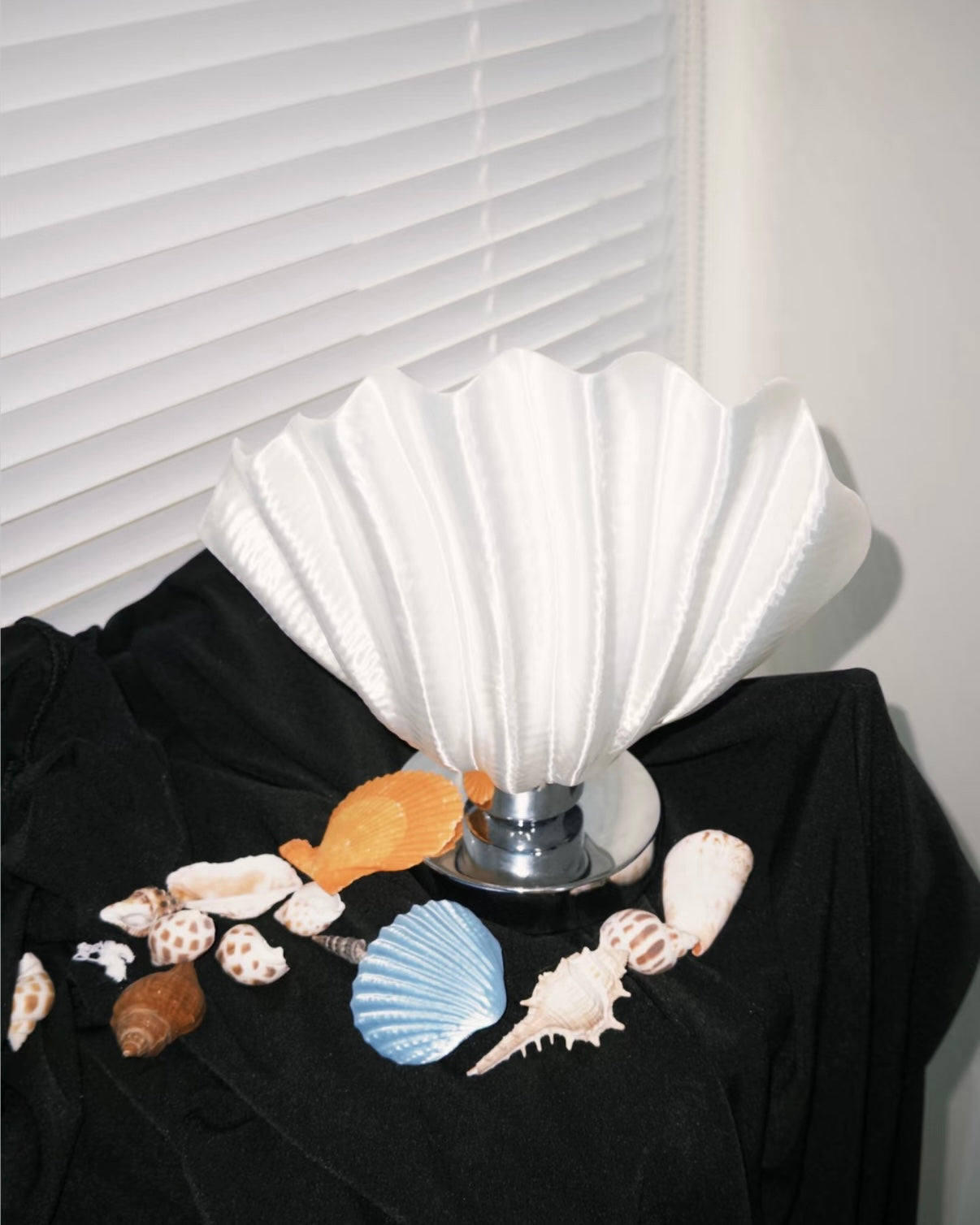 White shell-shaped lamp on a black surface with seashells around it, against a white wall.
