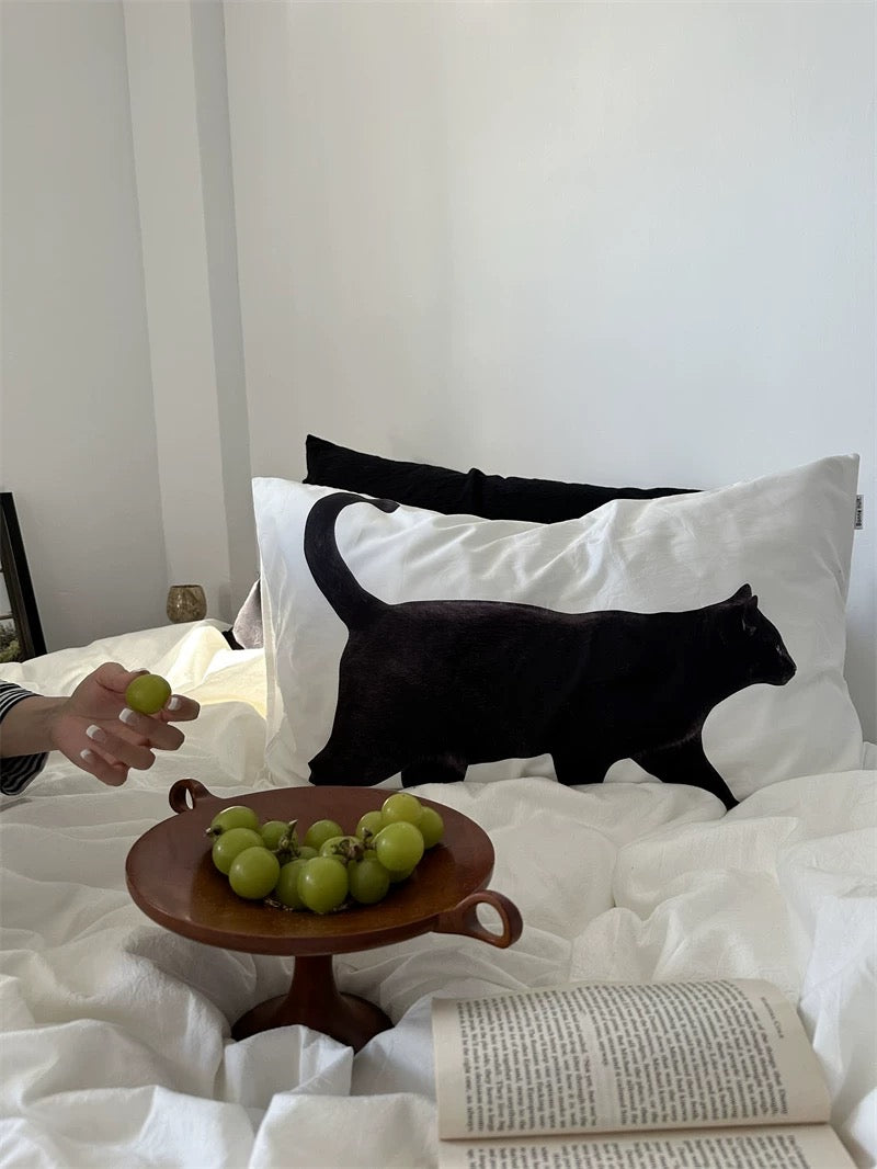 Cat-shaped pillow on a bed with a hand holding grapes and a book.