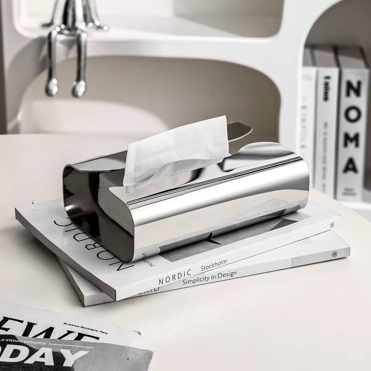 Silver tissue holder with tissues on top of a magazine in a minimalistic setting.