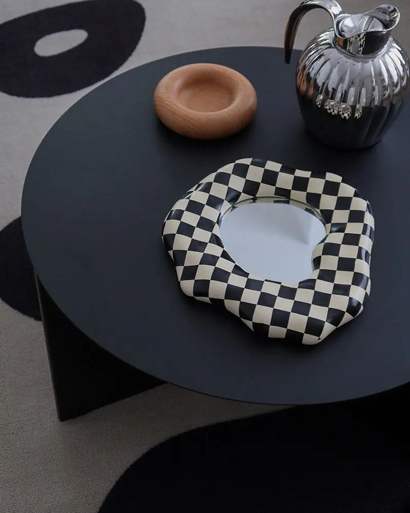 A scalloped-edge oval mirror with a black and cream checkerboard pattern, styled on a light wood cabinet next to a white ceramic vase and a basket with feathers.