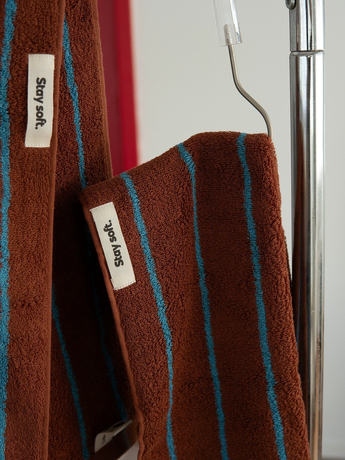 A close-up of a brown terrycloth towel with vertical turquoise blue stripes, hanging on a metal hanger from a silver metal stand. The brand name 'Stay soft.