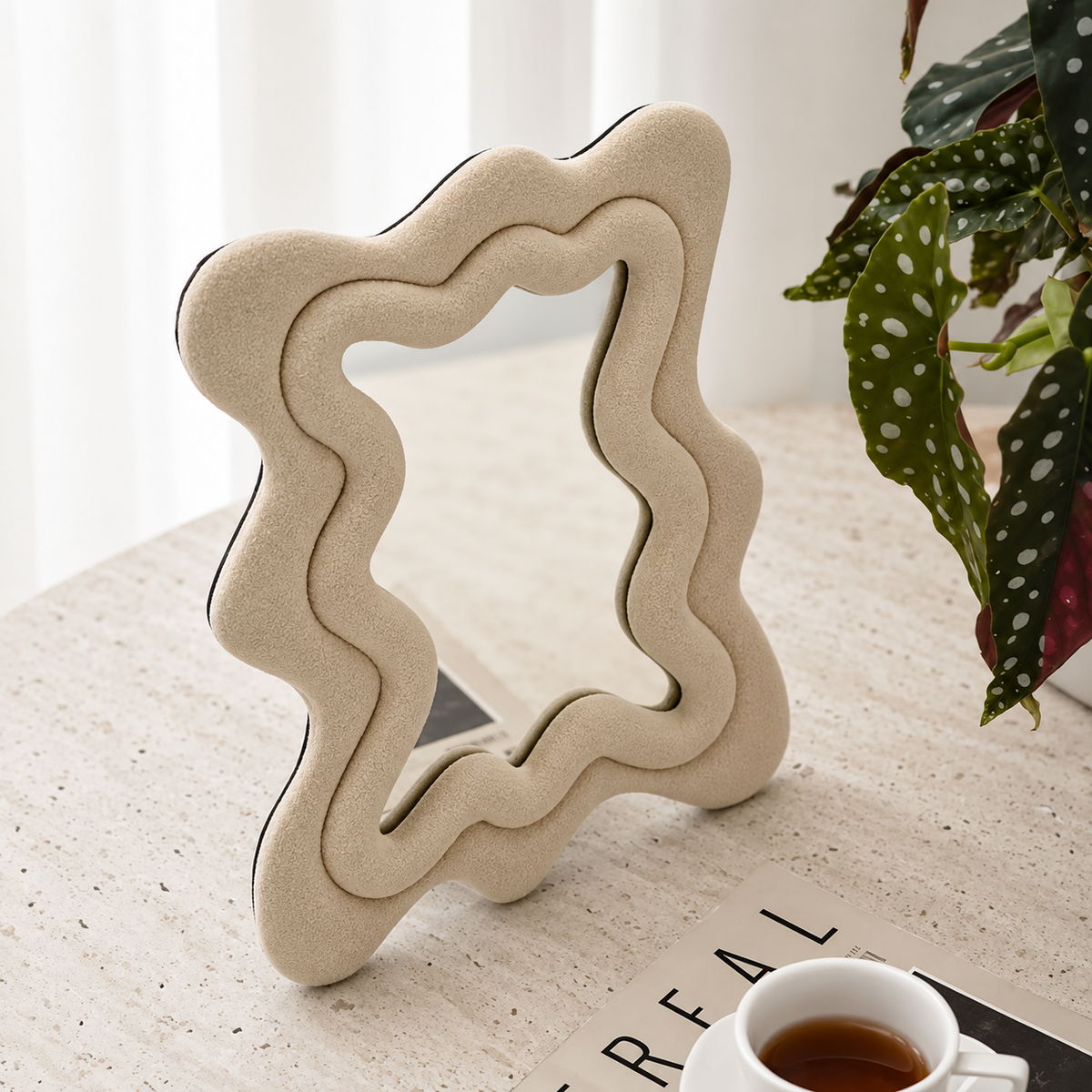 A wavy, star-shaped beige foam mirror stand with black trim, styled on a light stone tabletop next to a potted plant and a cup of tea.