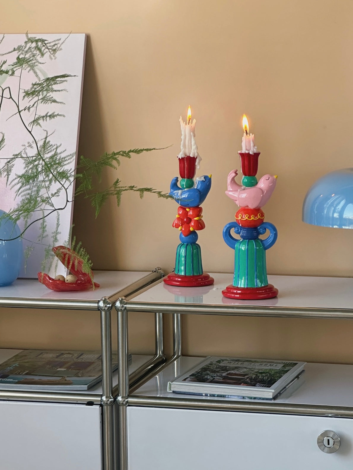 Pink and blue bird candle holders with lit candles styled beside stacked books on a mirrored console.
