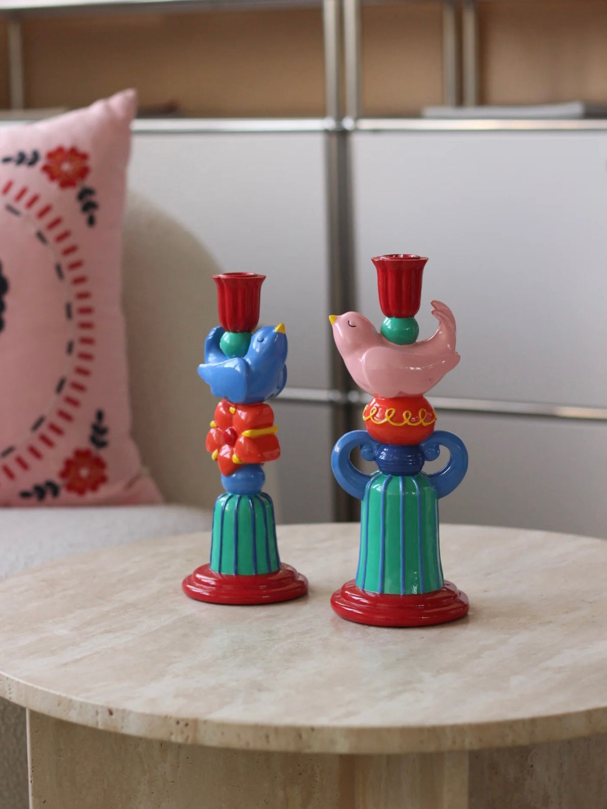 Pink and blue bird candle holders standing together on a round table with colorful decor in the background.