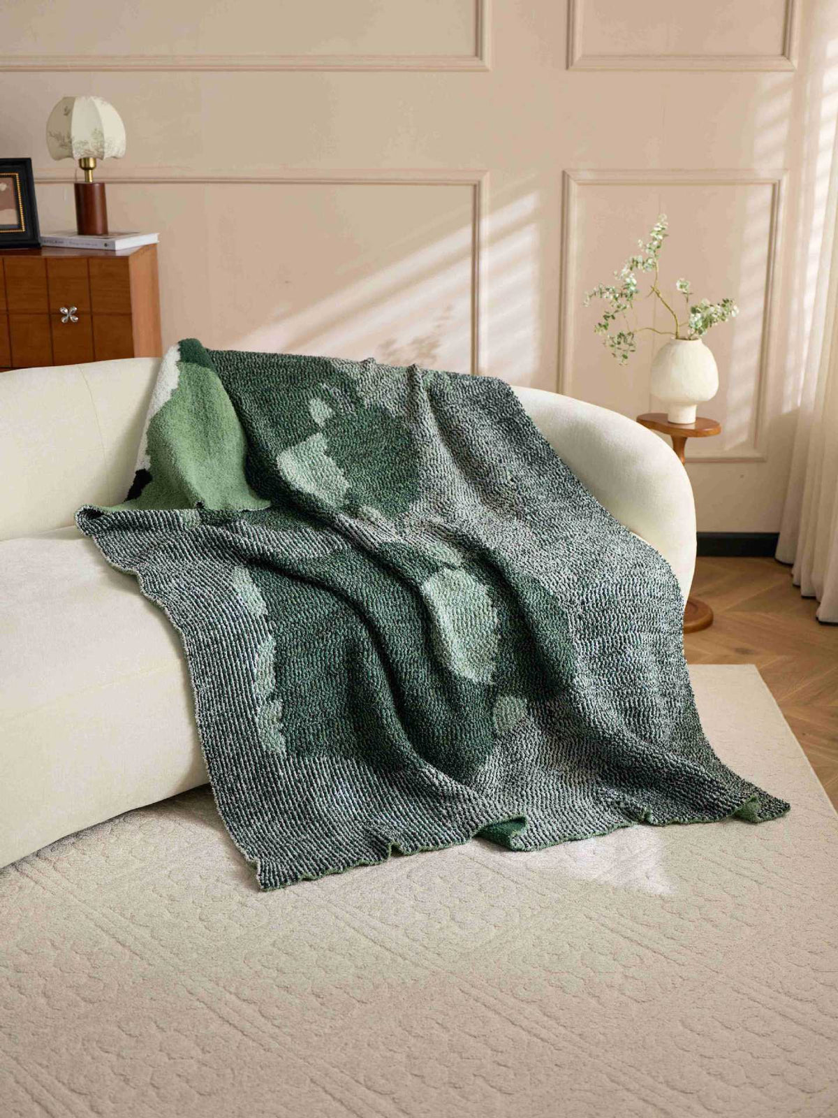 A textured knit throw blanket in shades of green and gray is draped over a white sofa in a softly lit living room.