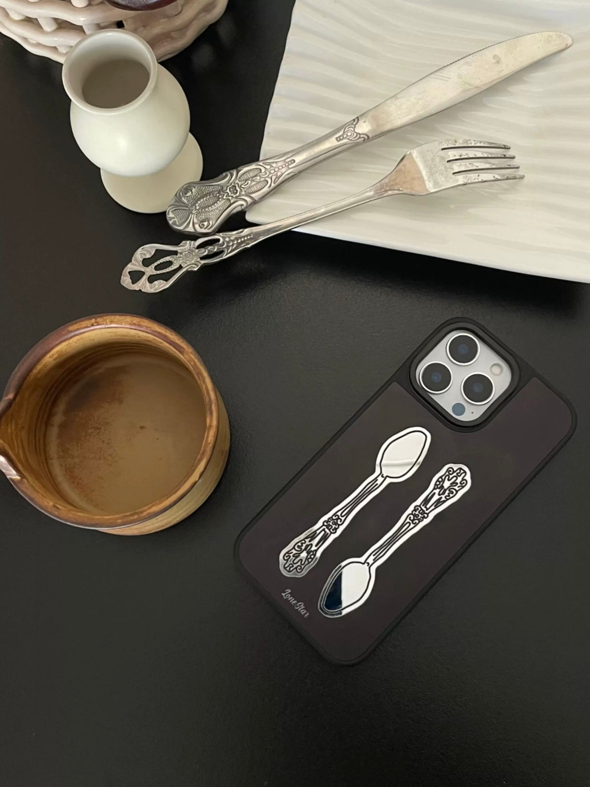 A black phone case lies diagonally with two silver colored vintage spoons facing in opposite directions.