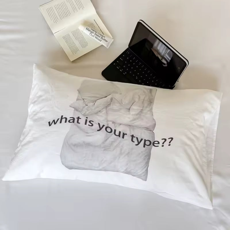 A white pillowcase laid flat on a bed, printed with a grayscale image of a rumpled sheet and the text 'what is your type??' in black. An open book and a black tablet with a keyboard rest nearby on the bedding.