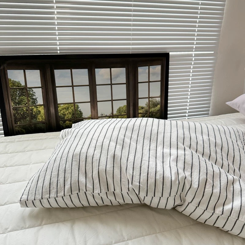 A white pillowcase with thin black stripes rests on white bedding beneath a framed window print.