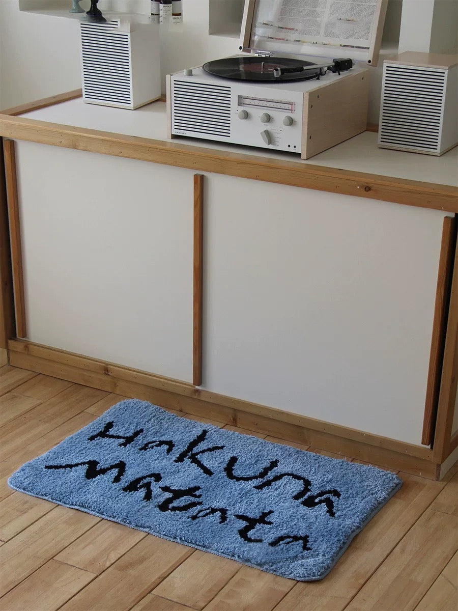 A blue bath mat with black lettering sits lengthwise in front of a cabinet on a pale indoor floor.