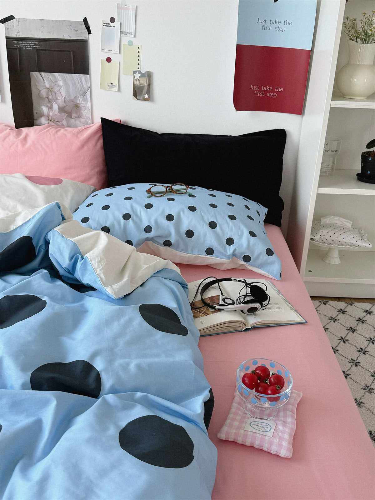 A small dotted blue pillow lies in front of a black pillowcase on a bed with a pink sheet and a blue dotted cover.