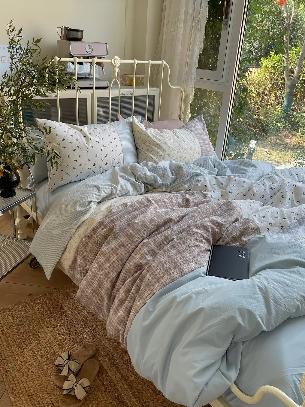 Blue and brown patchwork bedding spread across a cream metal bed beside a large window.