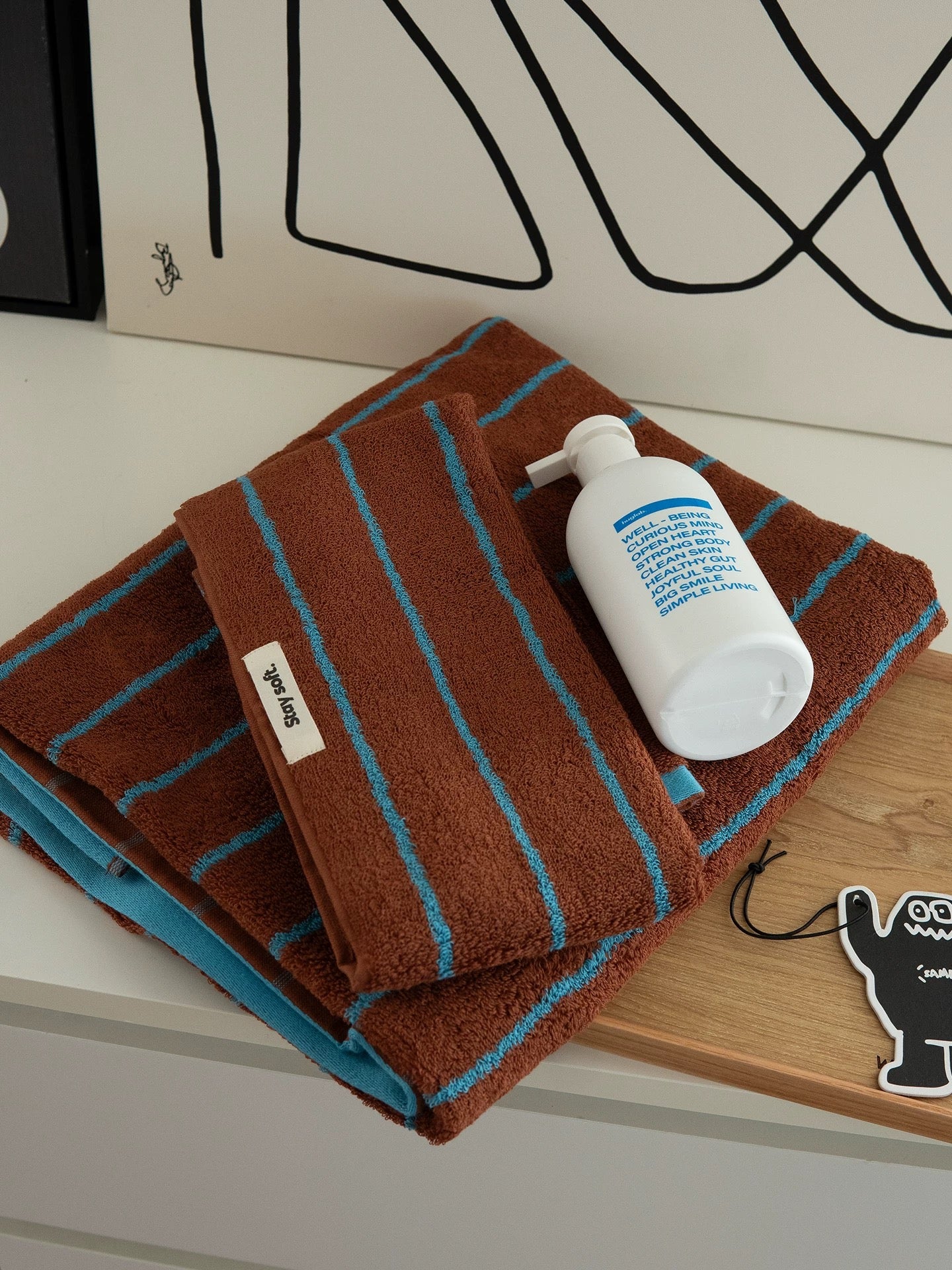 A stack of brown cotton towels with turquoise stripes, featuring a white pump bottle of hand wash and a small white brand tag reading 'sopapal' on a wooden tray.