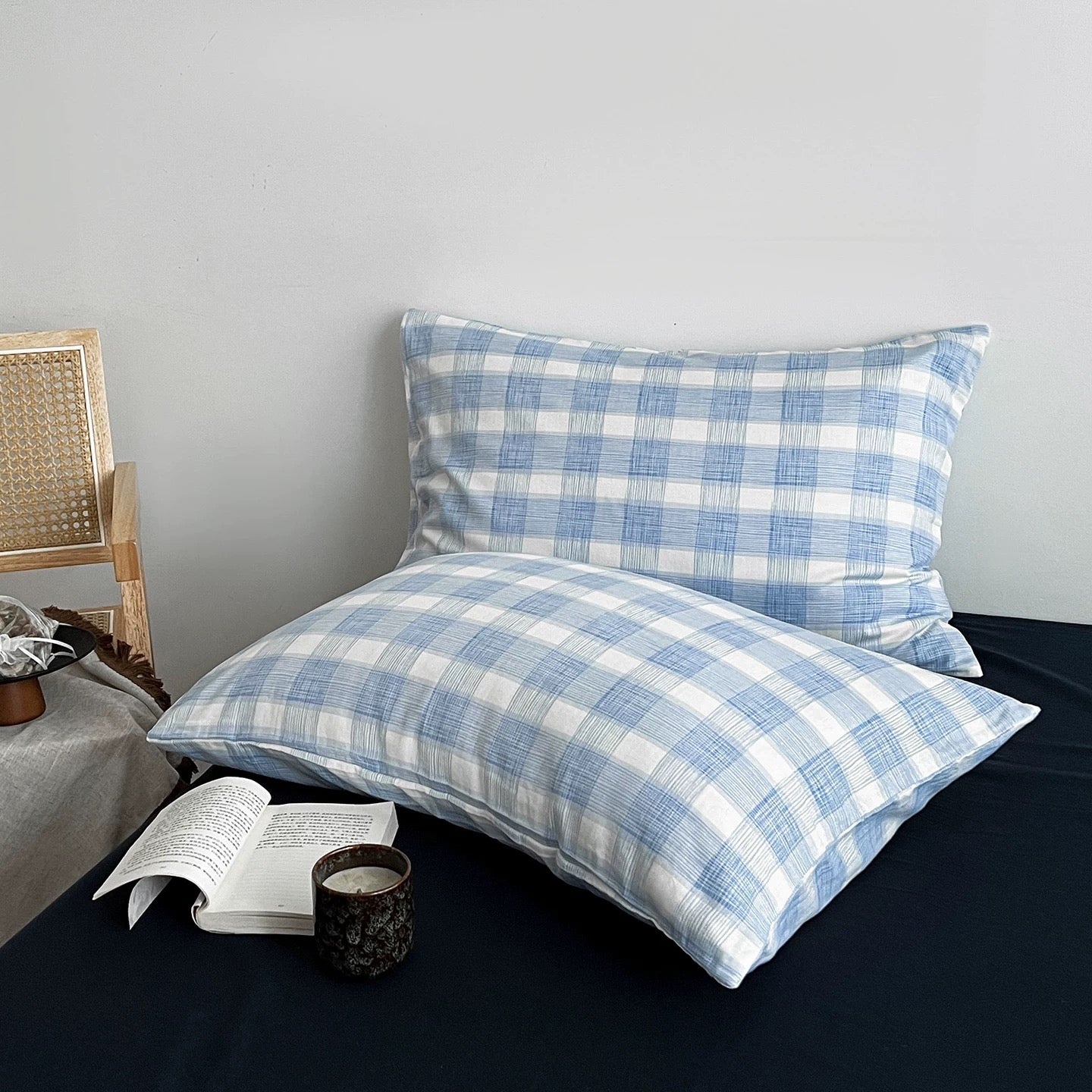 Two blue and white plaid pillowcases displayed on a bed, with an open book and a dark cup resting in front. A woven chair is partially visible on the left against a plain light gray wall.