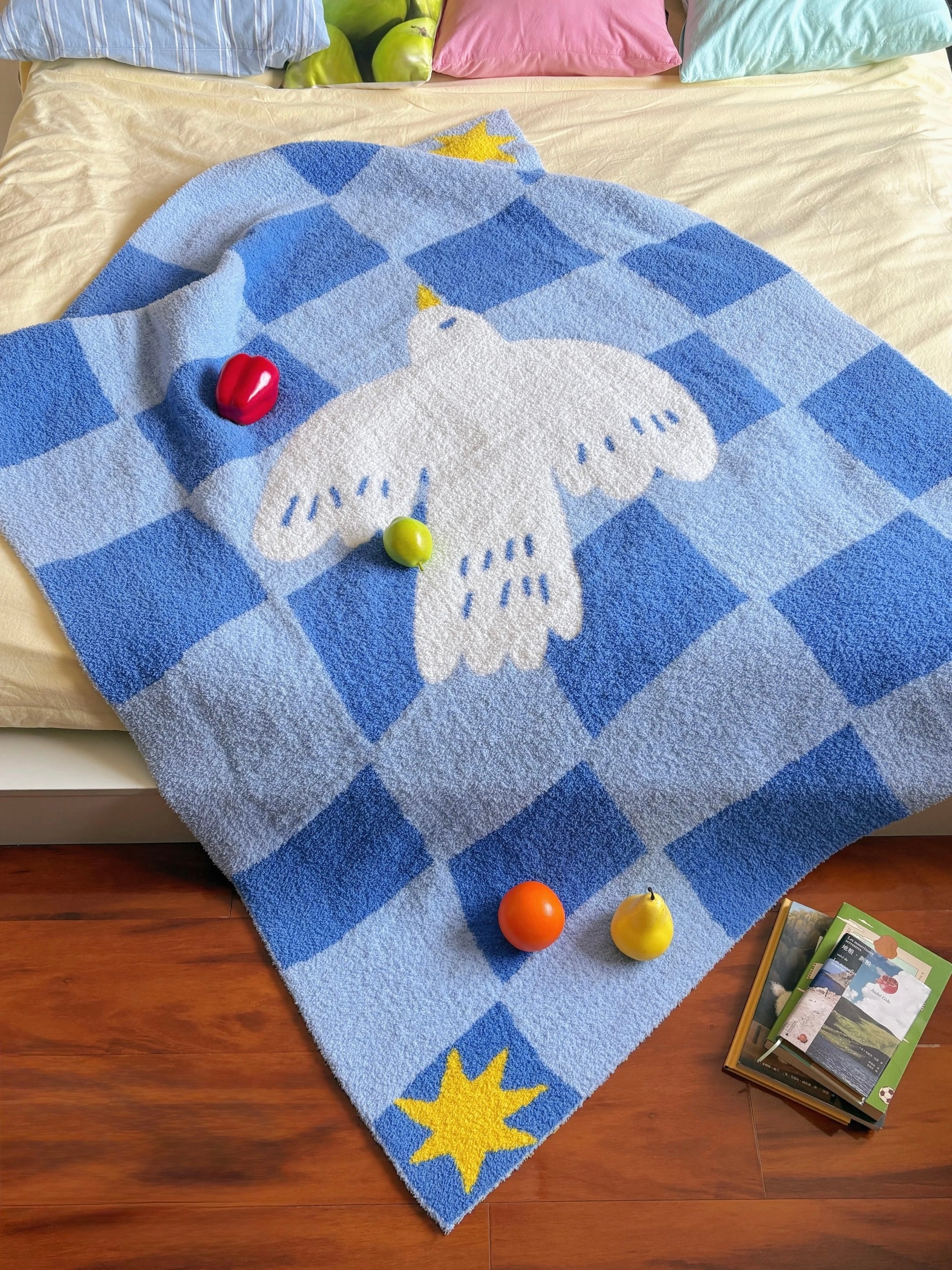 A blue and white checkered plush blanket with a white dove motif and yellow star accents, draped over a bed in a bedroom setting with colorful pillows, wooden floor, and small toys.