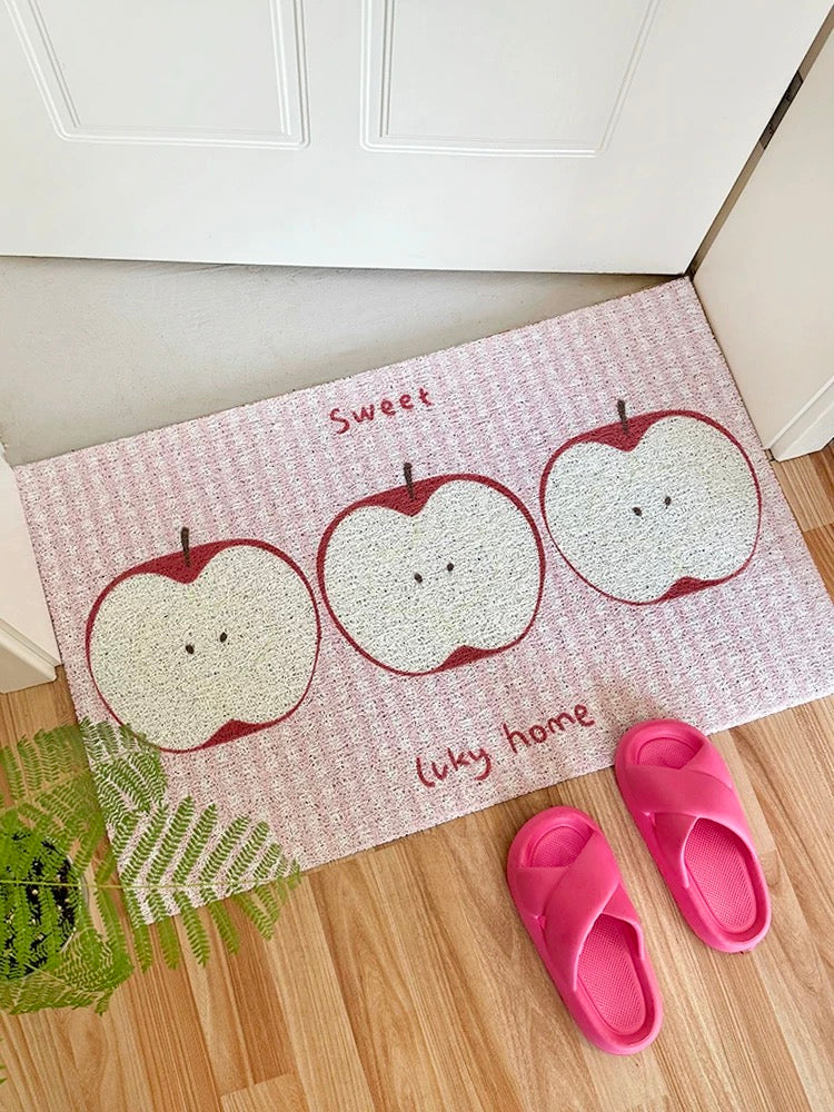 A rectangular entryway doormat with a pink and white textured background, featuring three cartoon apple illustrations and the words 'Sweet lucky home.' Pink slip-on sandals and a green plant are placed beside it on a wood floor in front of a white door.