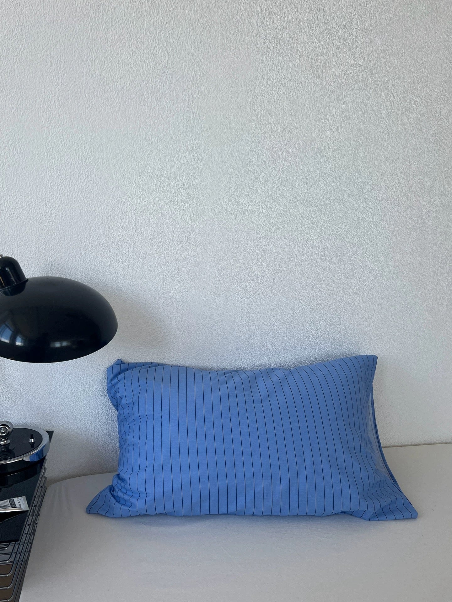 A rectangular blue pillow with thin vertical white pinstripes resting on a white surface against a white wall, next to a black desk lamp.