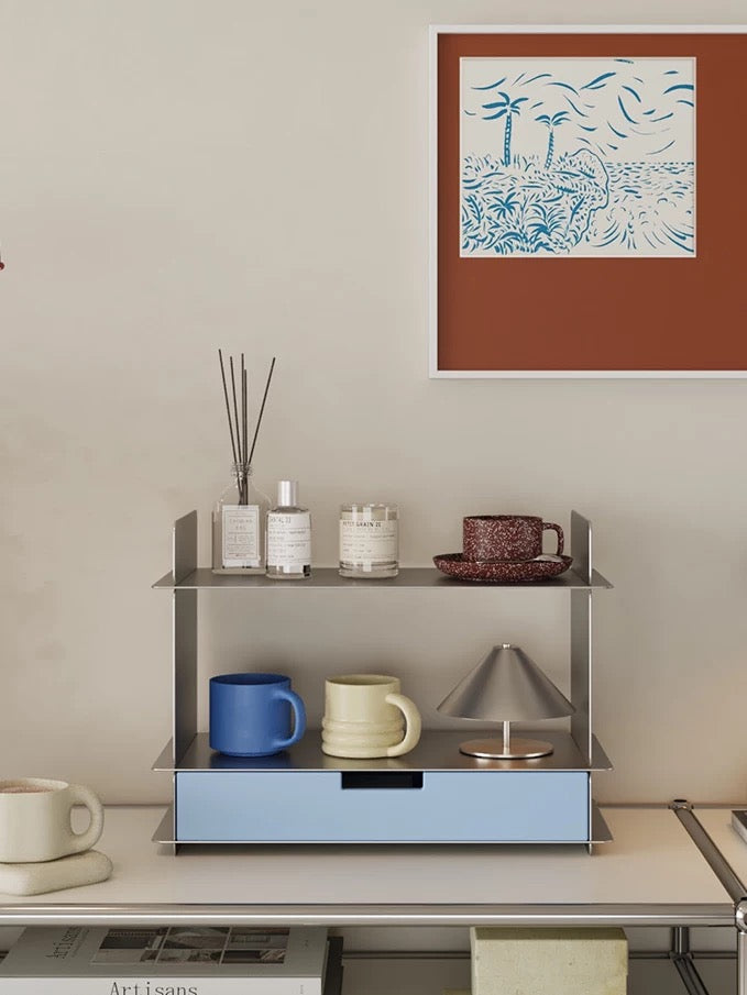 Blue desktop shelf with a pull out drawer, perfume bottles, a candle and mugs beside a framed palm tree print