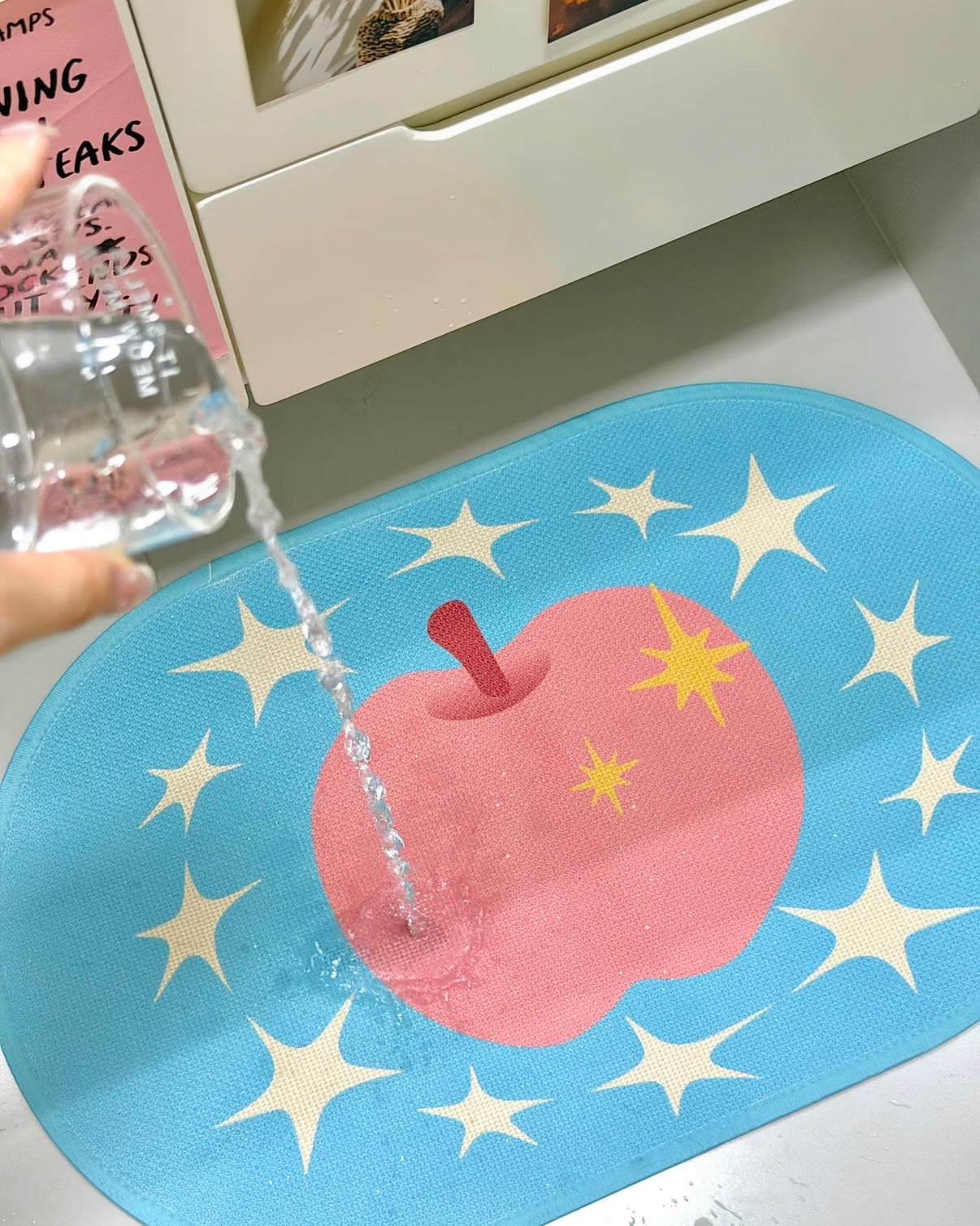A blue oval mat with a pink apple and yellow stars, shown with water being poured over it from a clear bottle. A hand is visible holding the bottle, and part of a white drawer or cabinet is in the background.