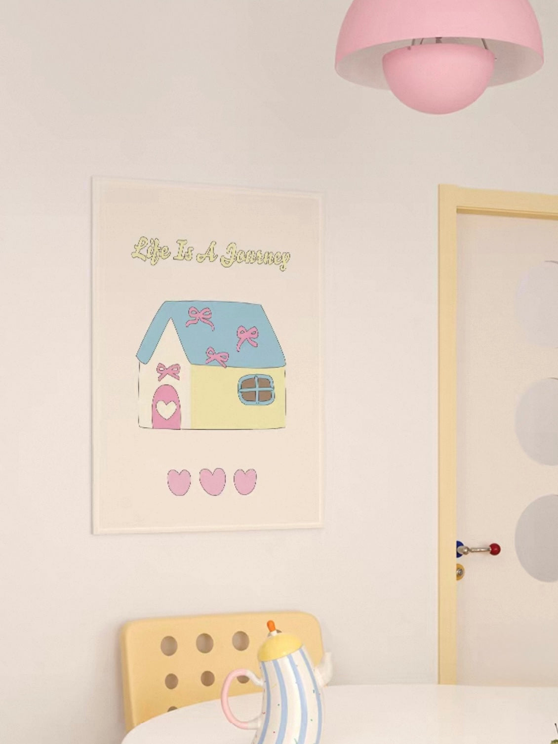A blue roof house tapestry shows a pale yellow home and pink hearts above a striped teapot on a table.