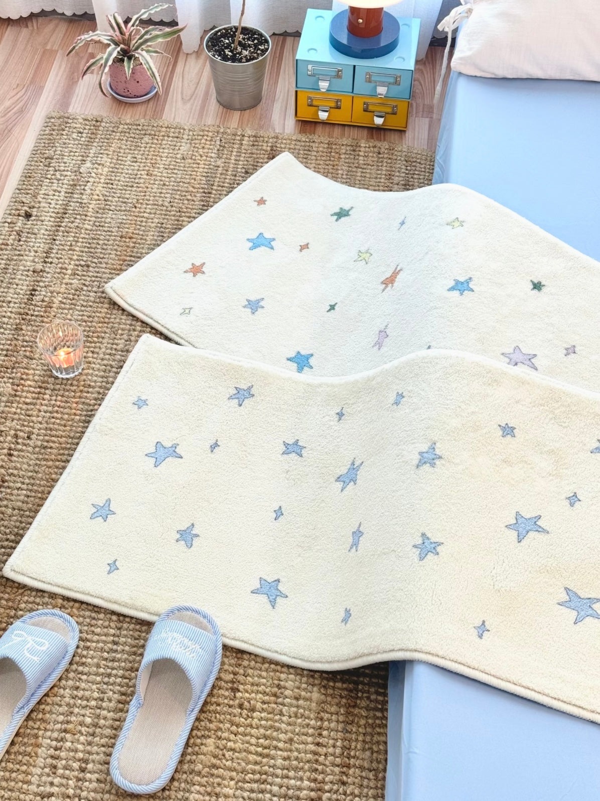 A cream rug with blue stars has a curved fold across its surface beside a blue bed and blue slippers.