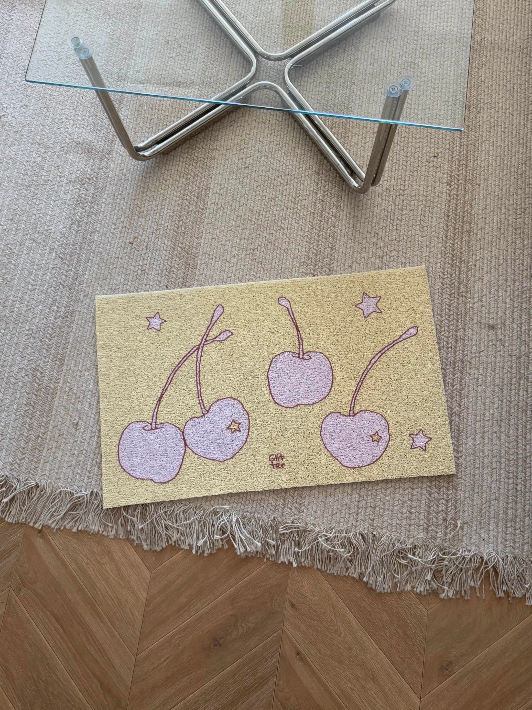 A rectangular decorative mat or tray with a pastel cherry illustration lies on a woven beige rug over a wood floor, with the lower legs of a glass-top table visible above.