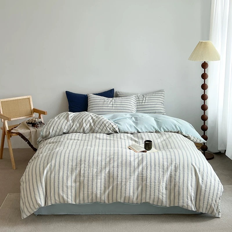 Two striped pillowcases stacked beside a navy pillow with an open book and ceramic mug