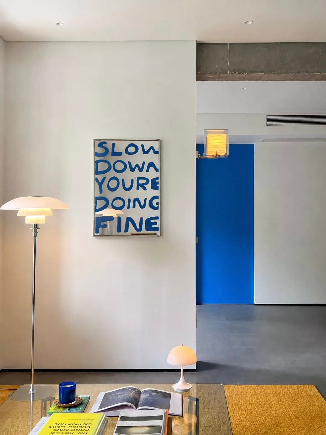 A blue text wall mirror hangs on a light wall, with stacked lettering and a slim silver rectangular frame.