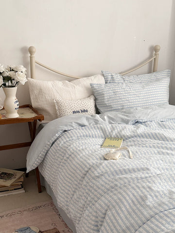 A neatly made bed with light blue and white striped bedding, including a duvet cover and matching pillowcases, styled in a cozy bedroom setting with a wooden side table, white flowers, and a small decorative pillow.