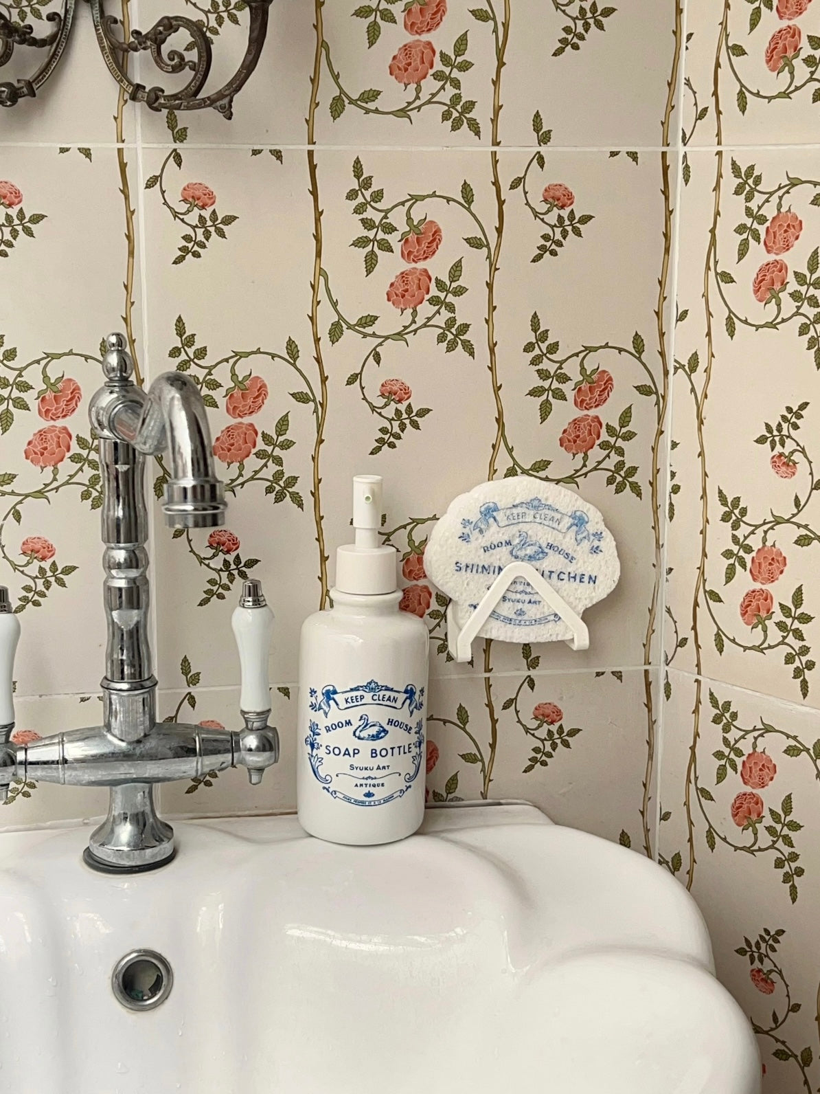 Decorative soap dispenser stands beside a faucet, with a matching scalloped sponge held above the sink.