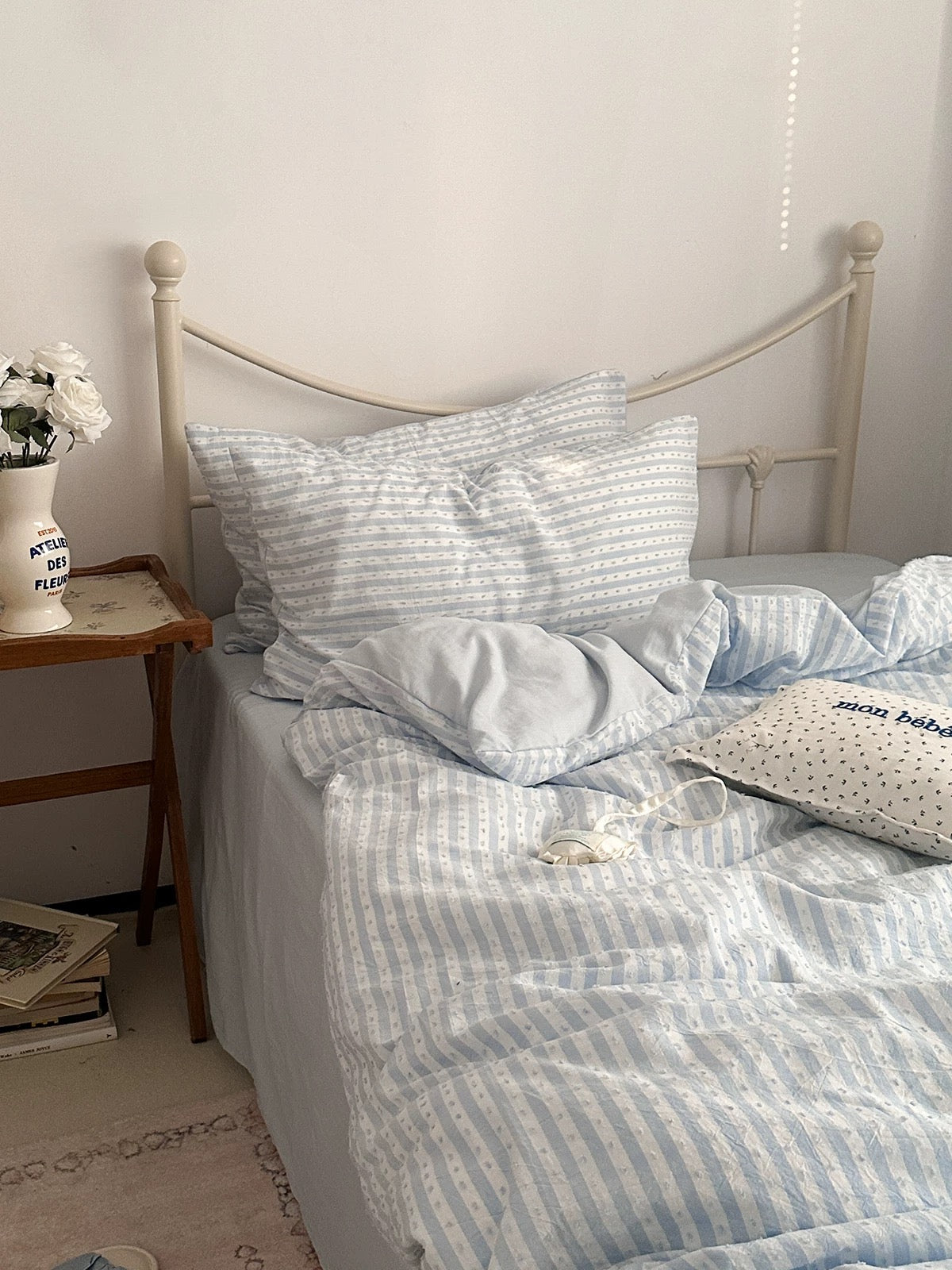 Light blue and white striped bedding set on a bed with a white metal headboard, styled in a cozy bedroom setting next to a wooden side table.