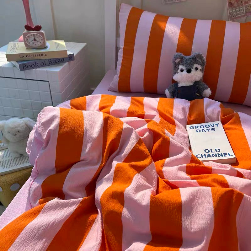 A cozy bed setup with orange and light pink striped bedding, including a duvet cover and pillowcase, styled with a small plush dog toy, books, and a bedside cabinet.
