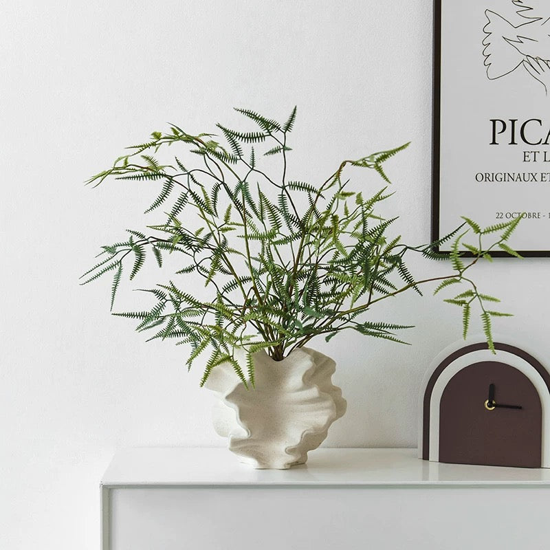 Decorative plant in a white vase on a shelf with a clock and framed picture in the background.