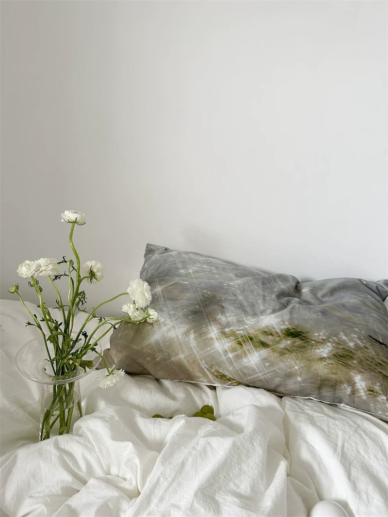 Gray pillow on a bed with white bedding and a vase of flowers against a light gray wall.