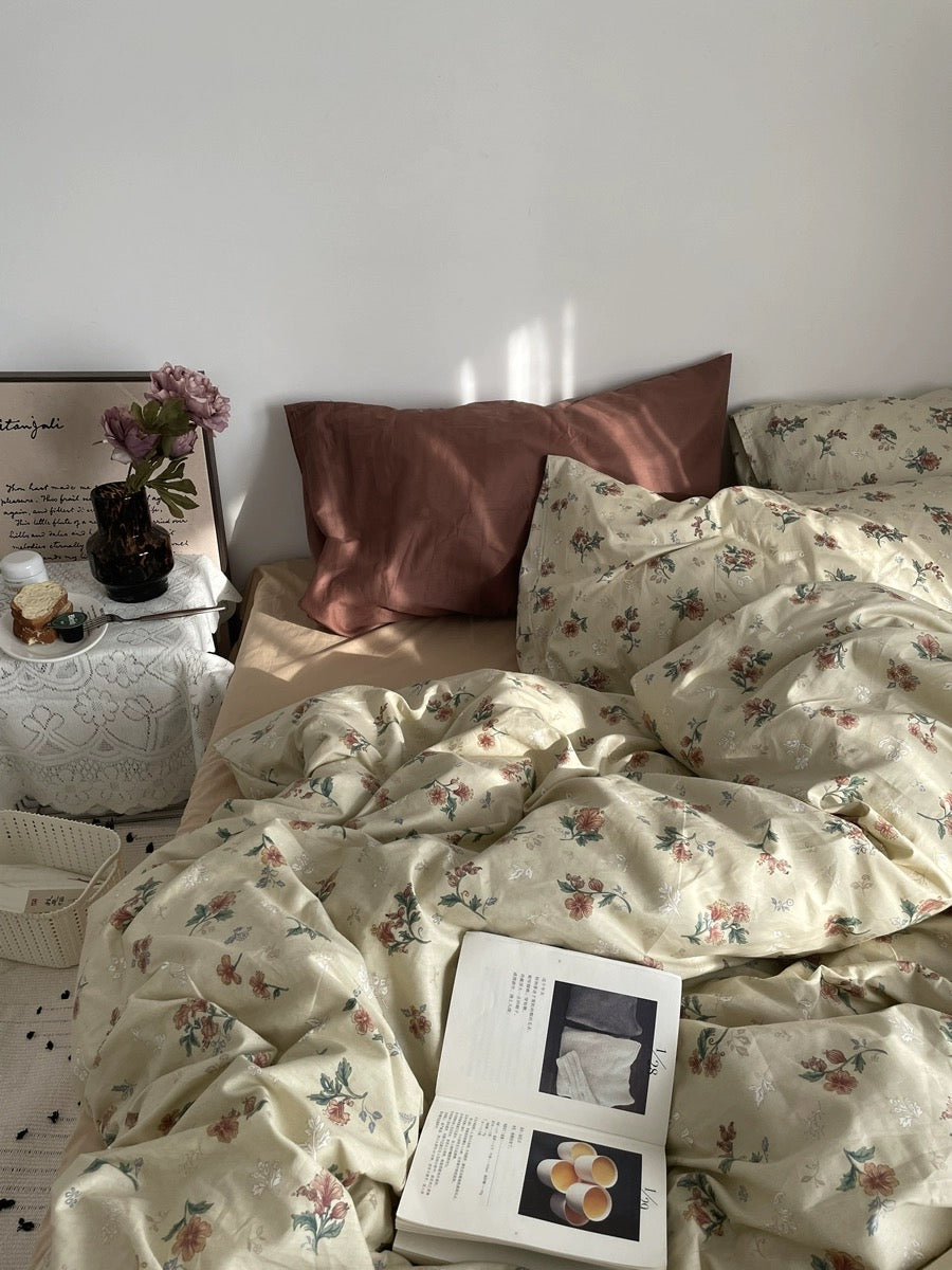 Botanical floral bedding styled on a sunlit bed with rust pillows, flowers, and an open book.