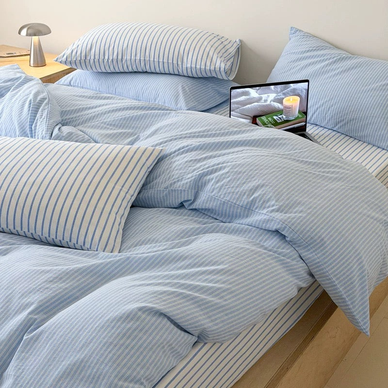 A light blue and white striped bedding set with matching pillows and duvet on a wooden platform bed, styled in a minimalist bedroom with a wooden nightstand and lamp.