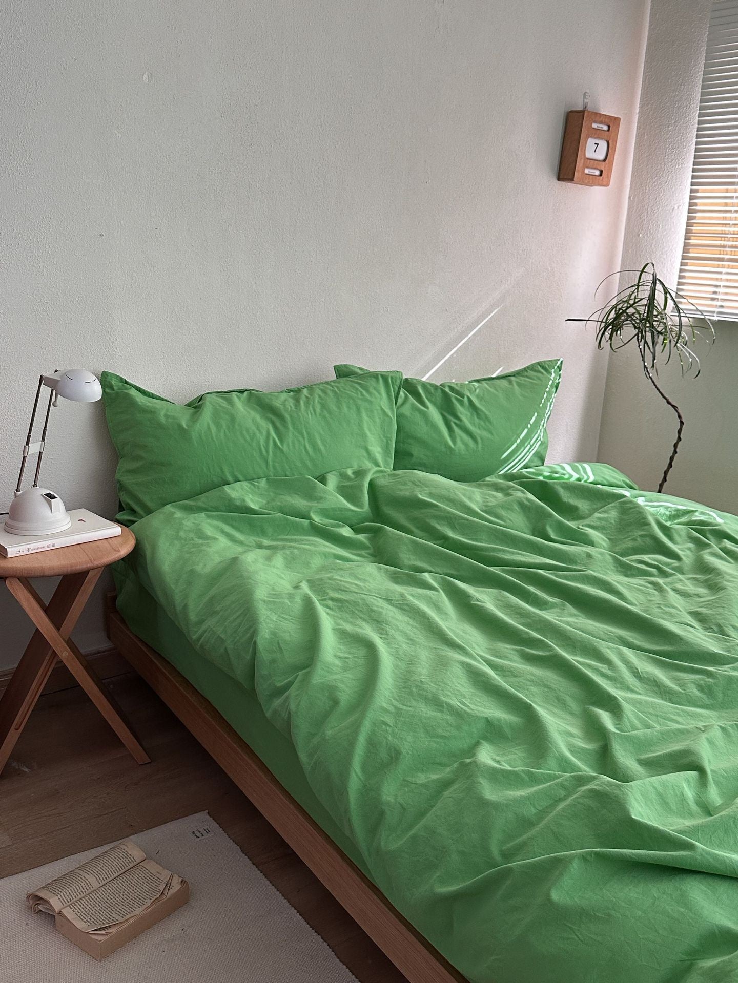 A bed dressed in bright green bedding, including two green pillows and a green duvet, styled in a minimalist bedroom with a wooden side table, white lamp, and a plant.