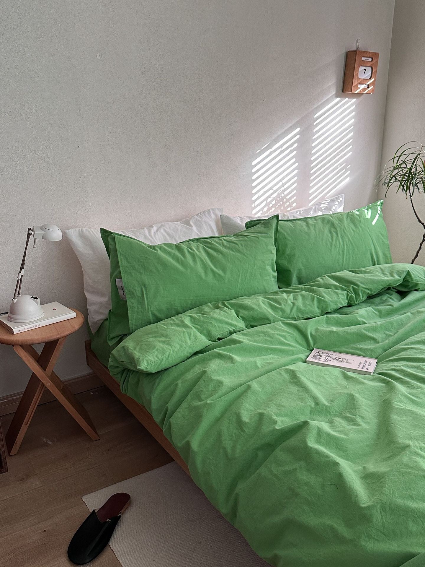 A bed dressed in a bright green bedding set with matching pillowcases and duvet, styled in a minimalist bedroom with a wooden side table, white lamp, and soft window light casting shadows.