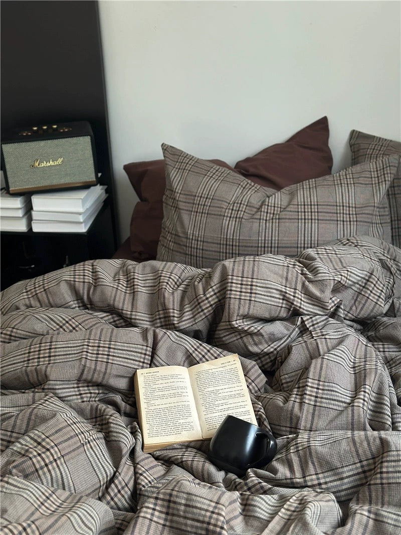 Bed with plaid comforter and pillows, featuring an open book and a mug.
