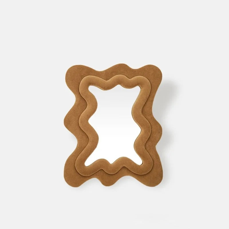 A decorative wall mirror with a wavy, layered frame in a warm tan or camel-colored suede-like material, shown against a plain light gray background.