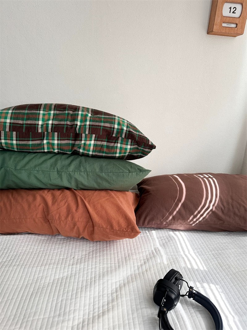 Plaid, green and rust orange pillowcases form a stack beside a cocoa brown design.