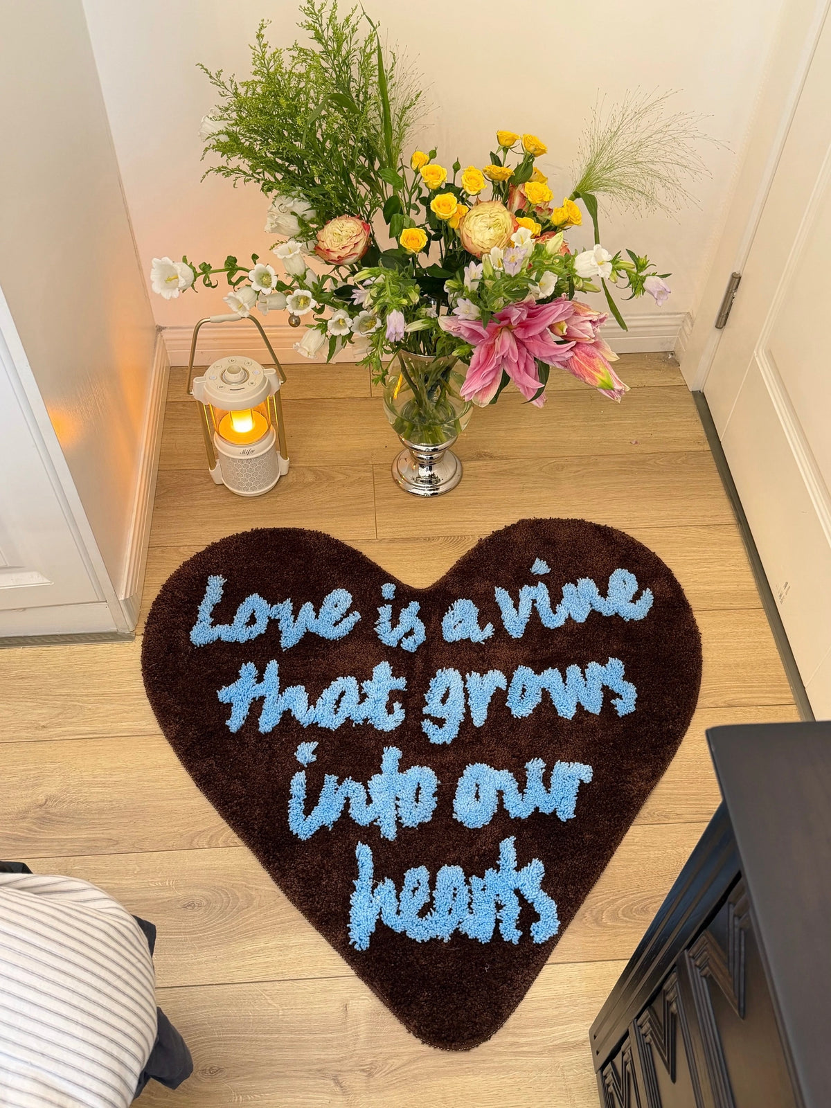 Brown heart rug with pale blue lettering sits near flowers and candles, with the full heart outline visible.